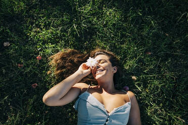 Smiling woman with eyes closed holding flower while relaxing on grassy land in park - DCRF00465