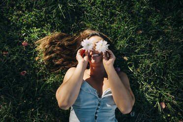 Young woman holding flowers over face while relaxing on grassy land in park - DCRF00466