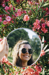 Nahaufnahme einer jungen Frau mit Sonnenbrille, die sich in einem Spiegel vor Blumen im Park spiegelt - DCRF00467