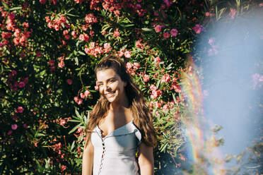Lächelnde junge Frau mit langen Haaren steht gegen Pflanzen im Park an einem sonnigen Tag - DCRF00469