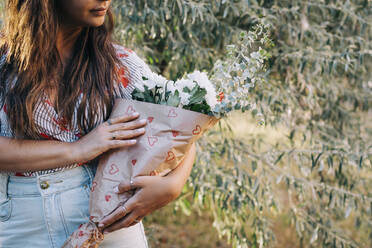 Nahaufnahme einer jungen Frau, die Blumen in der Hand hält, während sie im Park vor Pflanzen steht - DCRF00472