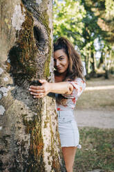 Lächelnde junge Frau, die einen Baumstamm hält, während sie im Park steht - DCRF00477