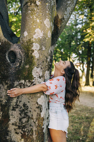 Cheerful young woman with long hair embracing tree trunk while standing in park stock photo