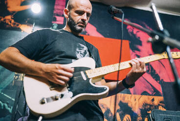 Blick von unten auf einen glatzköpfigen Mann im schwarzen T-Shirt, der auf der Bühne E-Gitarre spielt, vor dem Hintergrund von Fotos - ADSF02194