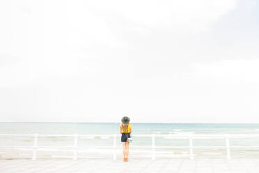 Rückansicht einer jungen Frau in elegantem Outfit, die in der Nähe eines Zauns am Ufer steht und den Blick auf das herrliche Meer genießt - ADSF02197