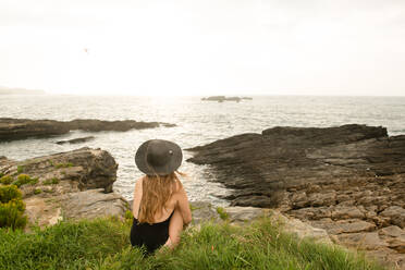 Rückenansicht einer jungen Frau mit Hut, die im Gras an der Küste sitzt und die Aussicht auf das herrliche Meer an einem sonnigen Tag bewundert - ADSF02198