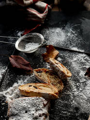 Scheiben von leckerem frischem Stollen liegen auf einer dunklen Tischplatte neben einem kleinen Sieb mit Puderzucker und trockenen Herbstblättern - ADSF02301