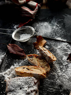 Scheiben von leckerem frischem Stollen liegen auf einer dunklen Tischplatte neben einem kleinen Sieb mit Puderzucker und trockenen Herbstblättern - ADSF02301