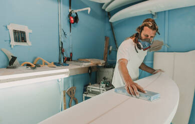 Mann in Atemschutzmaske mit Schleifpapier beim Polieren eines hölzernen Surfbretts am Arbeitsplatz - ADSF02325