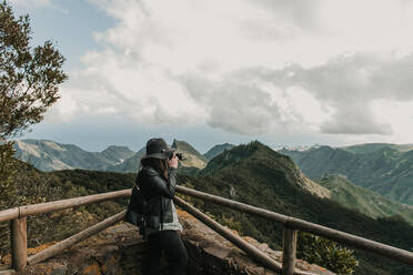 Side view of elegant lady in hat and leather jacket shooting on camera amazing view of peaks of hills and cloudy sky - ADSF02330