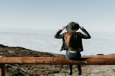 Rückansicht einer Touristin, die am Rande des Hügels auf einem Holzzaun sitzt - ADSF02340