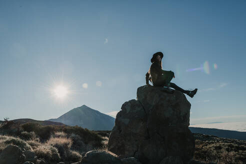 Seitenansicht einer nicht erkennbaren Reisenden, die auf einem Felsen am Hang sitzt, an einem sonnigen Tag - ADSF02342