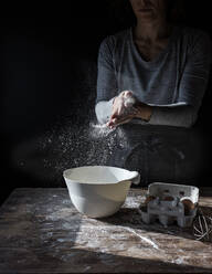 Crop lady clapping hands in flour near bowl, box of eggs and whisk on wooden table on black background - ADSF02452