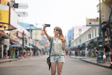 Fröhliche hübsche Frau, die ein Selfie macht und mit zwei Fingern am Strand von Khao San Road, Thailand, gestikuliert. - ADSF02476
