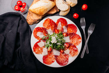 Leckere dünne Scheiben von ruckartigem Fleisch, bestreut mit gehackten Tomaten und Kichererbsen, auf einem weißen Keramikteller mit Gabeln auf einem schwarzen Holztisch von oben - ADSF02557