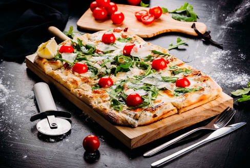 Auf einem Holzbrett servierte, frische, rechteckig geschnittene Pizza mit Kirschtomaten, Erdbeeren und Käse auf einem dunklen Tisch, auf dem Mehl, Gemüse, Gabel und Pizzaschneider von oben gestreut sind - ADSF02559