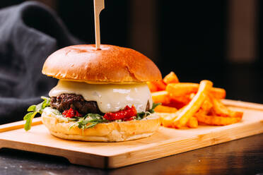 Nahaufnahme eines frischen Burgers mit Fleischpastete und Gemüse auf einem Holzbrett mit leckeren Pommes frites auf einem dunklen Holztisch - ADSF02564