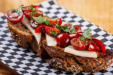 Close-up view of slice of rye bread with thin slices of cheese and halves of fresh cherry tomatoes placed on checkered board on wooden table - ADSF02569