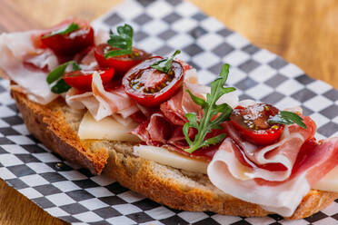 Close-up view of piece of bread with slices of cheese, delicious bacon, halves of cherry tomatoes and potherbs placed on checkered board on wooden table - ADSF02571