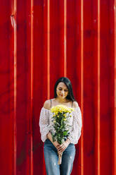 Young woman holding flowers while standing against red wall - DCRF00482