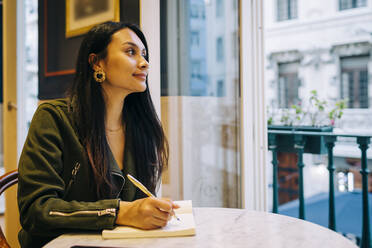 Lächelnde junge Frau, die im Café sitzend in ein Buch schreibt - DCRF00491