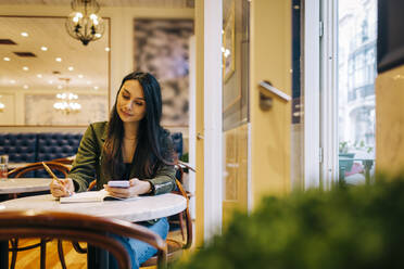 Lächelnde junge Frau, die in einem Café in ein Buch schreibt und telefoniert - DCRF00493