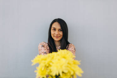 Young woman offering flowers while standing against wall - DCRF00498