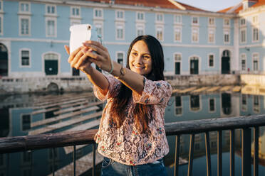 Young woman taking selfie through camera while standing on bridge - DCRF00500