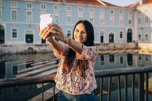 Junge Frau, die ein Selfie durch die Kamera macht, während sie auf einer Brücke steht - DCRF00500