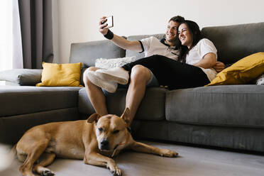 Paar macht Selfie auf Sofa in der Nähe von Hund zu Hause - EGAF00432