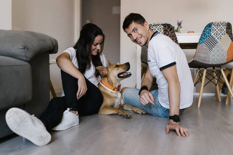 Lächelndes Paar sitzt mit Hund zu Hause auf dem Boden, lizenzfreies Stockfoto