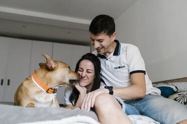 Dog kissing woman by man on bed at home - EGAF00458