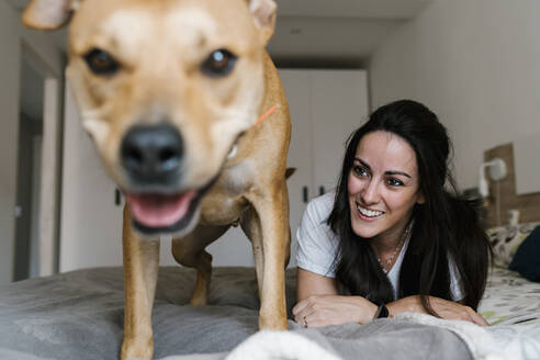Lächelnde Frau sieht ihren Hund an, während sie zu Hause auf dem Bett liegt - EGAF00459