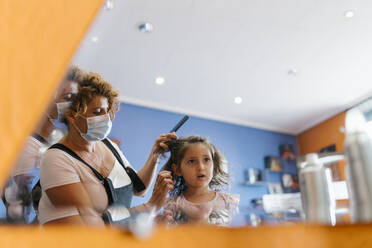 Mature female hairdresser cutting girl's hair seen from mirror reflection at barber shop - EGAF00473