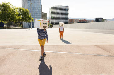 Boys wearing robot masks posing on road in city during sunny day - VABF03133