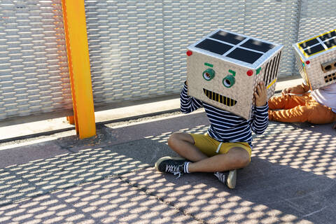 Boys wearing robot masks made of boxes sitting on road by fence in city stock photo
