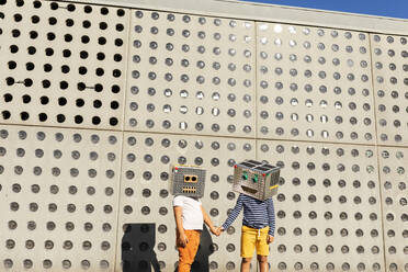 Friends wearing robot costumes holding hands while standing against wall on sunny day - VABF03141