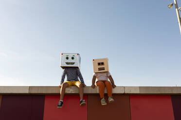 Friends wearing robot masks sitting on retaining wall against clear blue sky - VABF03142