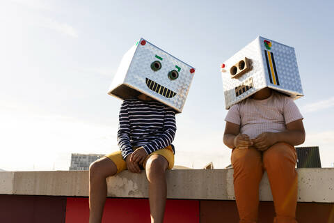 Freunde mit Robotermasken sitzen auf einer Stützmauer gegen den klaren Himmel an einem sonnigen Tag, lizenzfreies Stockfoto