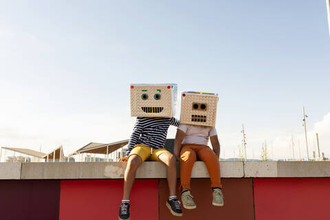 Freunde in Roboterkostümen sitzen im Sommer auf einer Stützmauer gegen den klaren Himmel, lizenzfreies Stockfoto