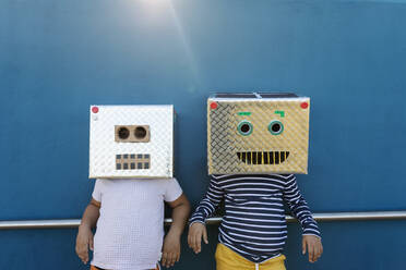 Friends wearing robot costumes made of boxes while standing against blue wall - VABF03149