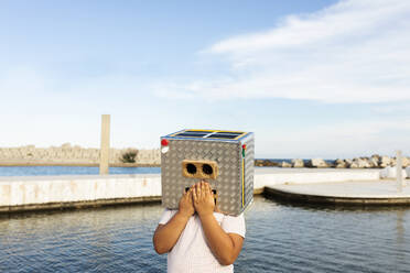 Junge mit Händen auf dem Mund der Robotermaske am Wasser stehend gegen den Himmel - VABF03151