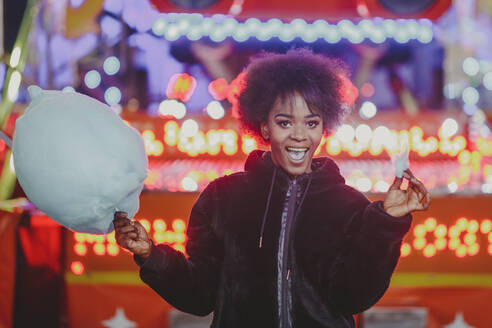 Attraktive afroamerikanische Frau mit leckerer Zuckerwatte, die lächelt und in die Kamera schaut, während sie in einem nächtlichen Vergnügungspark steht - ADSF02646