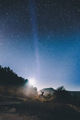 Seitenansicht in der Ferne einer Frau, die am Straßenrand unter blauem Sternenhimmel mit hellem Licht im Hintergrund steht - ADSF02666