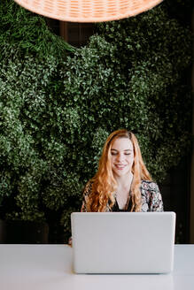 Cheerful pretty redhead woman sitting at laptop and typing at the table. - ADSF02697