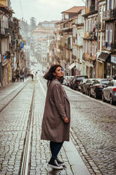 Seitenansicht einer hübschen jungen Dame in einem eleganten Mantel, die wegschaut, während sie auf einer gepflasterten Straße in einer alten Stadt steht - ADSF02776