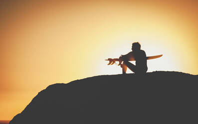 Seitenansicht von männlichen Surfer dunkle Figur sitzt auf einem Hügel und hält Brett in den Händen auf Hintergrund beleuchtet bei Sonnenuntergang - ADSF02783