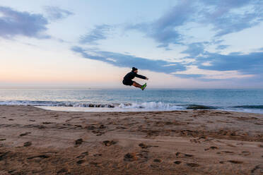 Handsome man in sportswear jumping high during outdoor training on sandy beach - ADSF02870