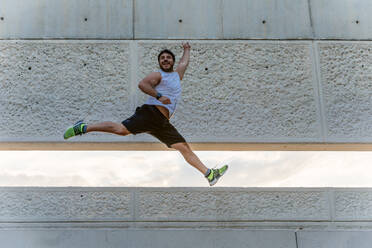 Attractive bearded man in sportswear smiling and jumping high during outdoor training on street - ADSF02873