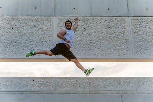 Attractive bearded man in sportswear smiling and jumping high during outdoor training on street - ADSF02873